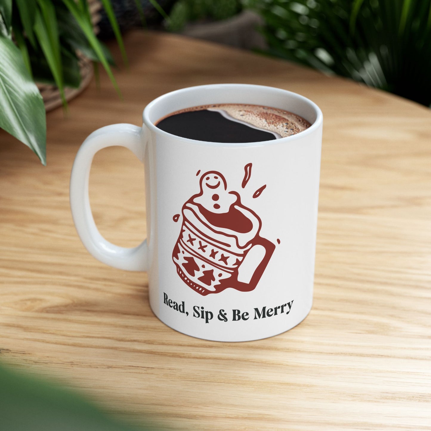 White mug with a red illustration and text 'Read, Sip & Be Merry' on a wooden surface.