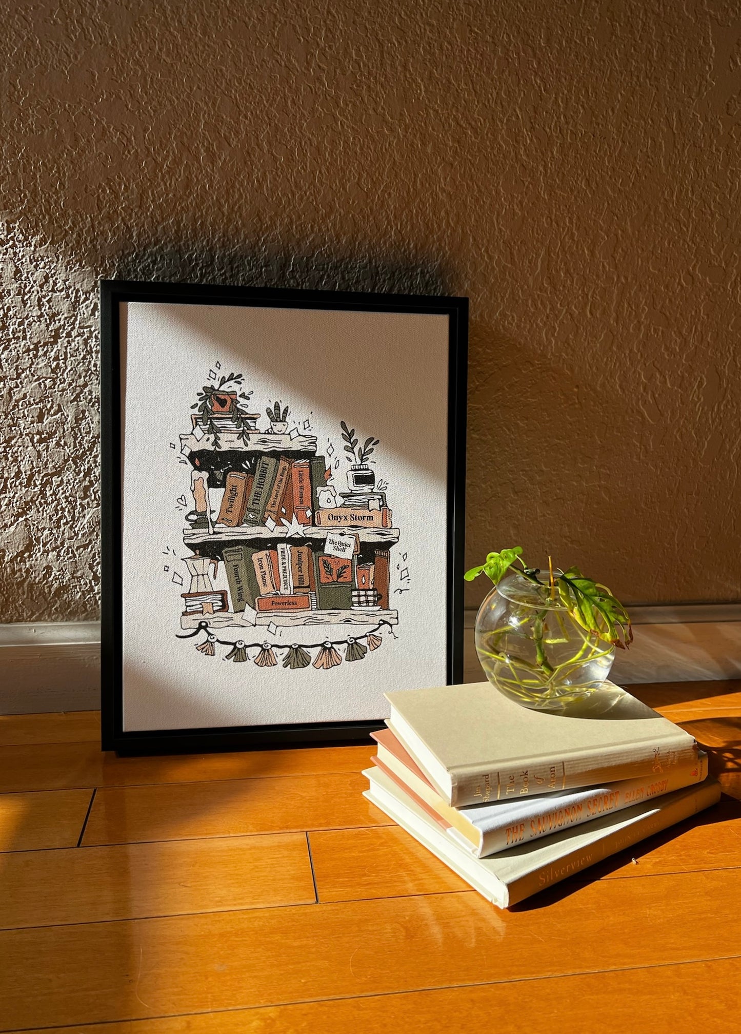 Framed artwork of a bookshelf on a wooden floor with books and a plant.