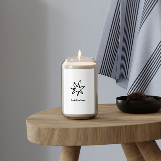 A white candle with a black star design and the text 'Booked and busy' on the label, with a lit wick, placed in a clear glass jar with a metal lid on top of a wood stool