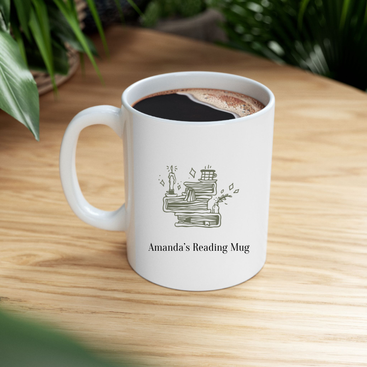 A white ceramic mug with a personalized name 'Amanda's Reading Mug' printed on the front, featuring a quiet shelf icon and book illustrations, on top of a wooden table