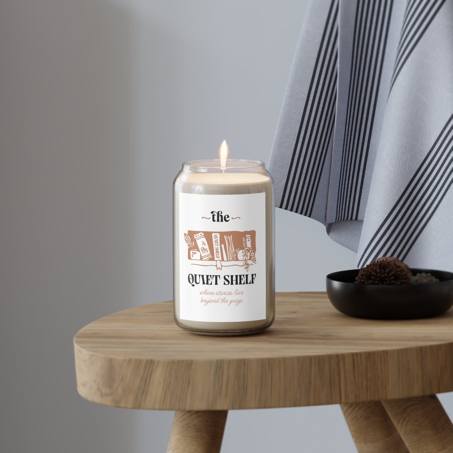A soy candle in a clear glass jar with a minimalist label that features a book and the text 'the QUIET SHELF', with a lit wick, on a wood stool