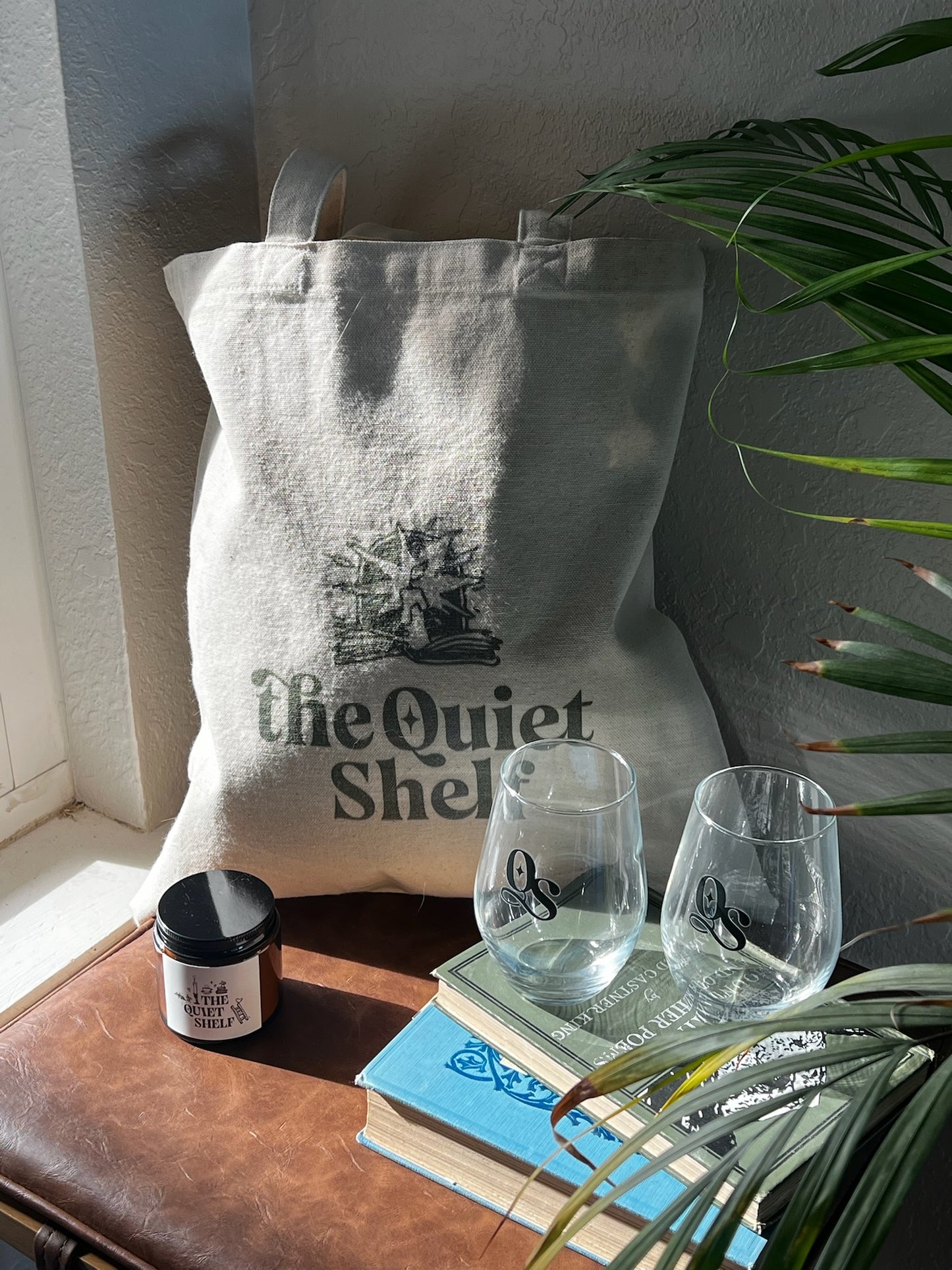 Tote bag with 'the Quiet Shelf' branding on a surface with books, wine glasses, and a candle.