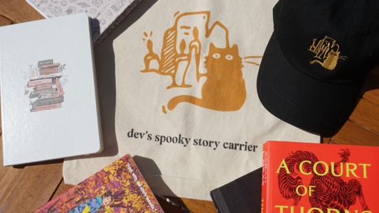 Flat lay of personalized bookish gifts including a canvas tote bag reading “dev’s spooky story carrier” with a cat and candles design, a black embroidered hat, a book stack journal, and fantasy novels.