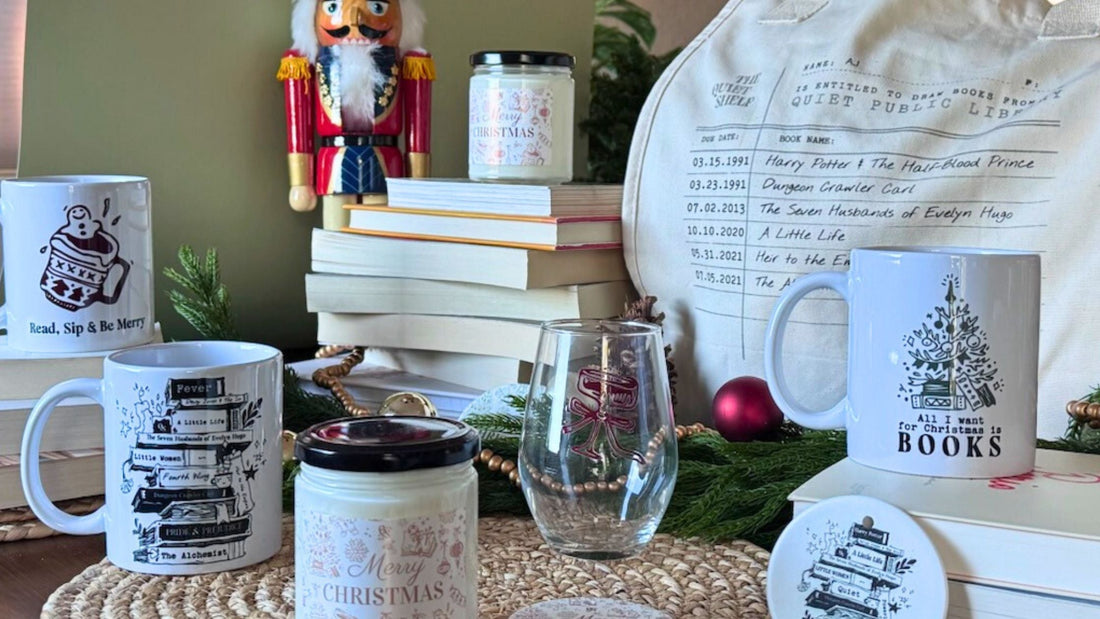 A festive tabletop scene with bookish Christmas mugs, a personalized library card tote bag, candles, stemless wine glass, ornament, and stacked books, styled with a nutcracker figurine, greenery, and red ornaments.