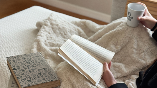 Reader relaxing on the couch with a blanket, holding a Quiet Shelf mug and reading a book.