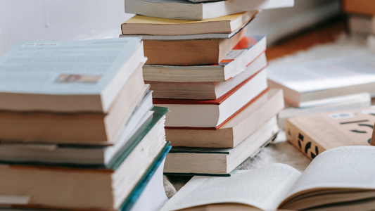 Stacks of paperback and hardcover books piled high with an open book in the foreground, representing a towering TBR pile.