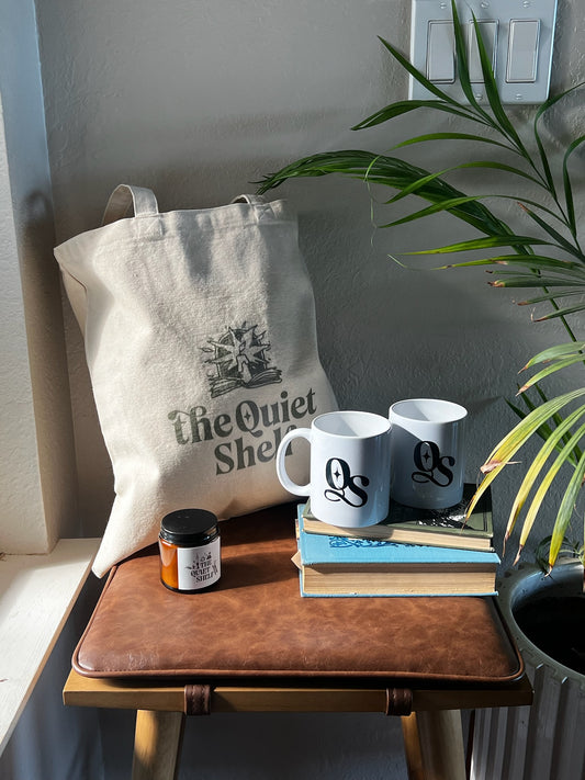 Tote bag with 'The Quiet Shop' branding, two mugs, a candle, and books on a wooden surface.