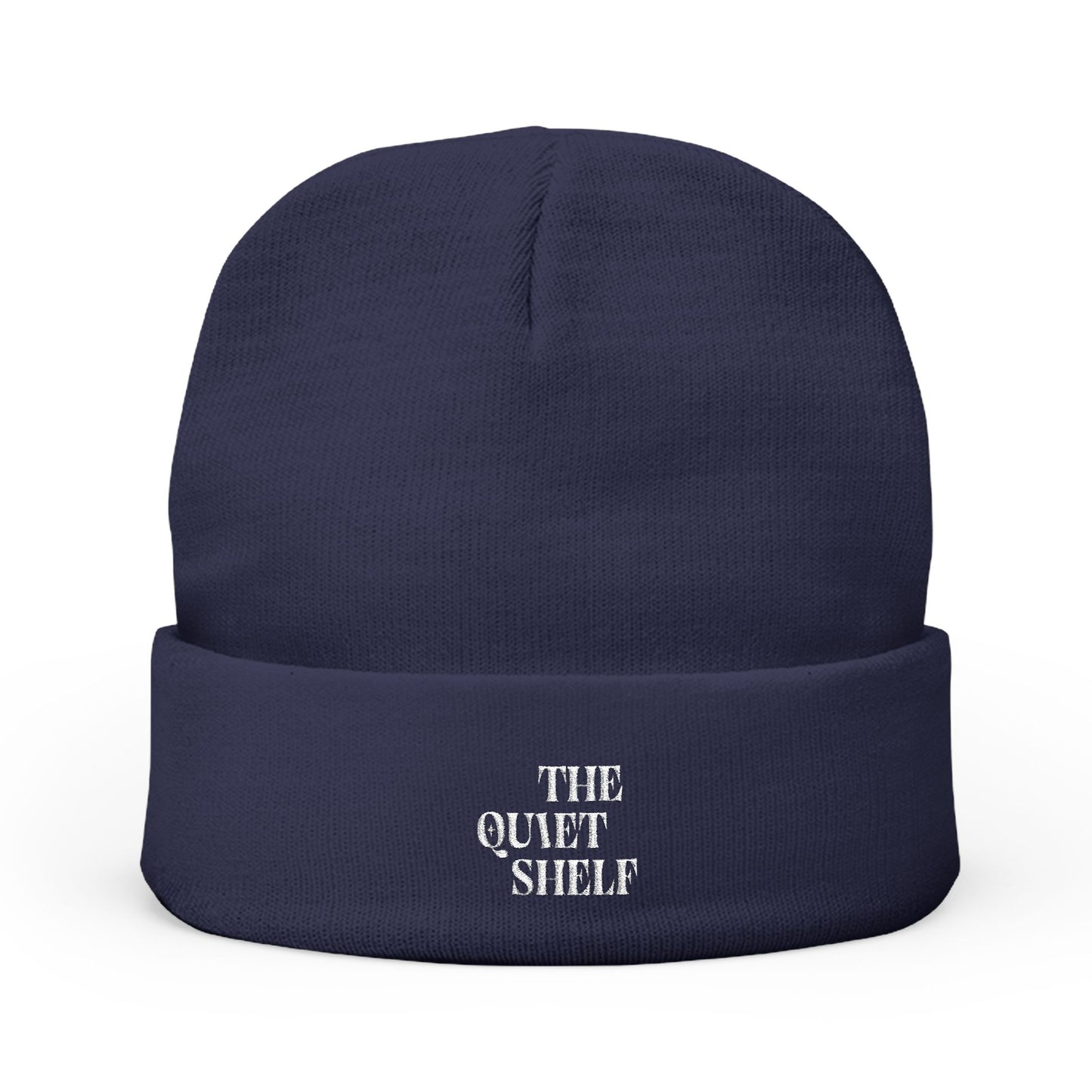 A navy blue cuffed beanie with 'THE QUIET SHELF' embroidered in white on the fold-over cuff.