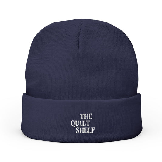 A navy blue cuffed beanie with 'THE QUIET SHELF' embroidered in white on the fold-over cuff.