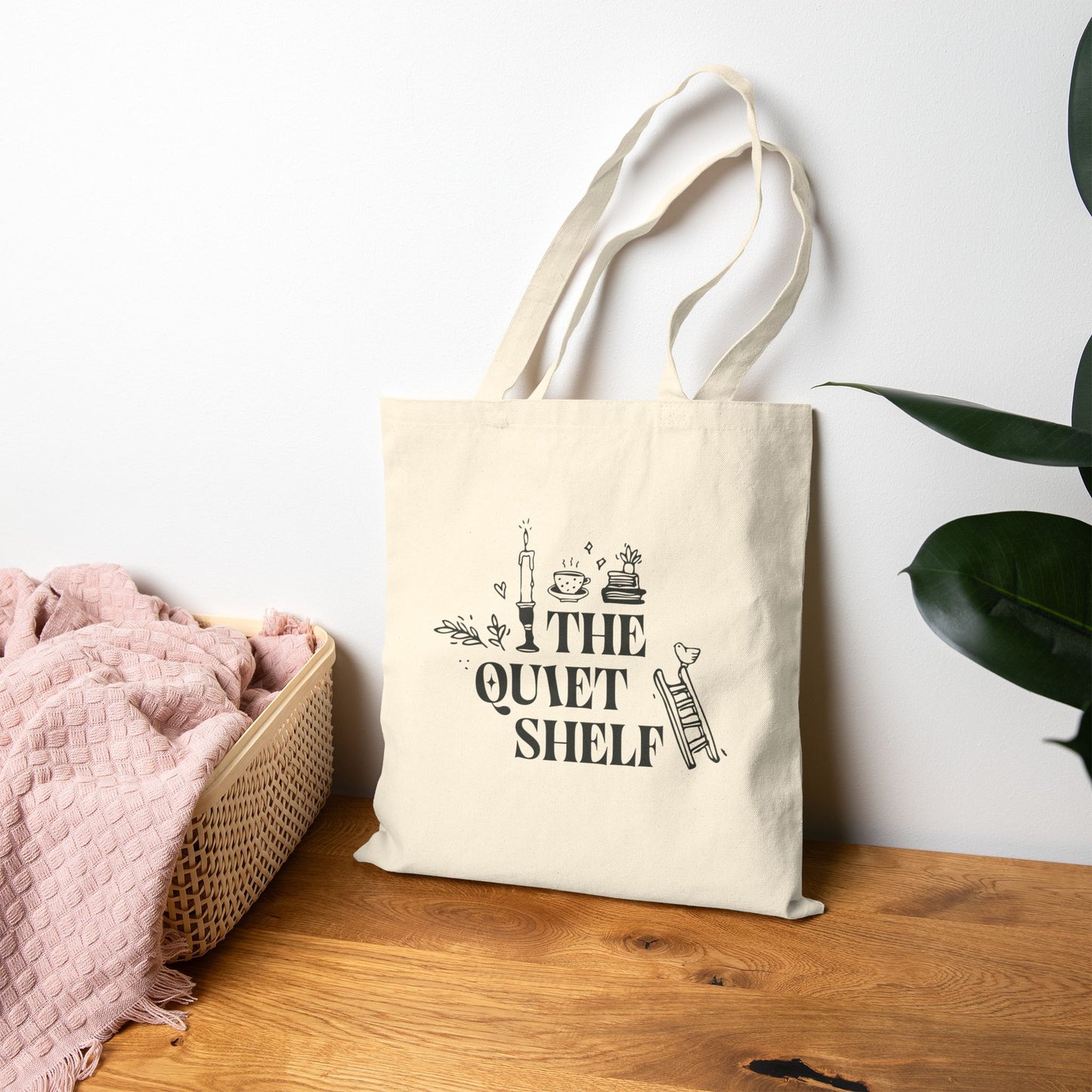 Beige tote bag with 'The Quiet Shelf' design on a wooden surface with a plant in the background