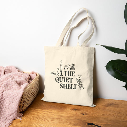 Beige tote bag with 'The Quiet Shelf' design on a wooden surface with a plant in the background