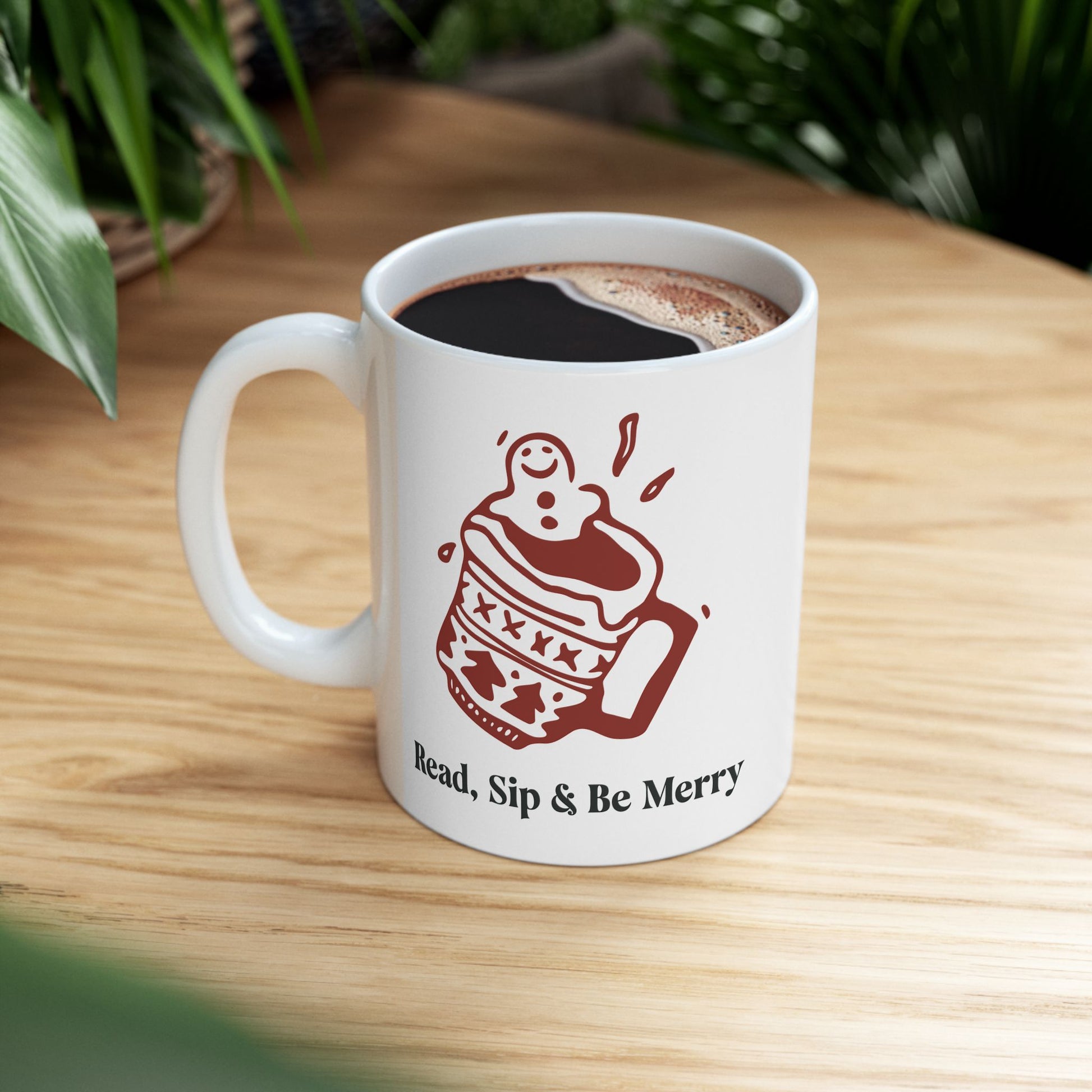 White mug with a red illustration and text 'Read, Sip & Be Merry' on a wooden surface.