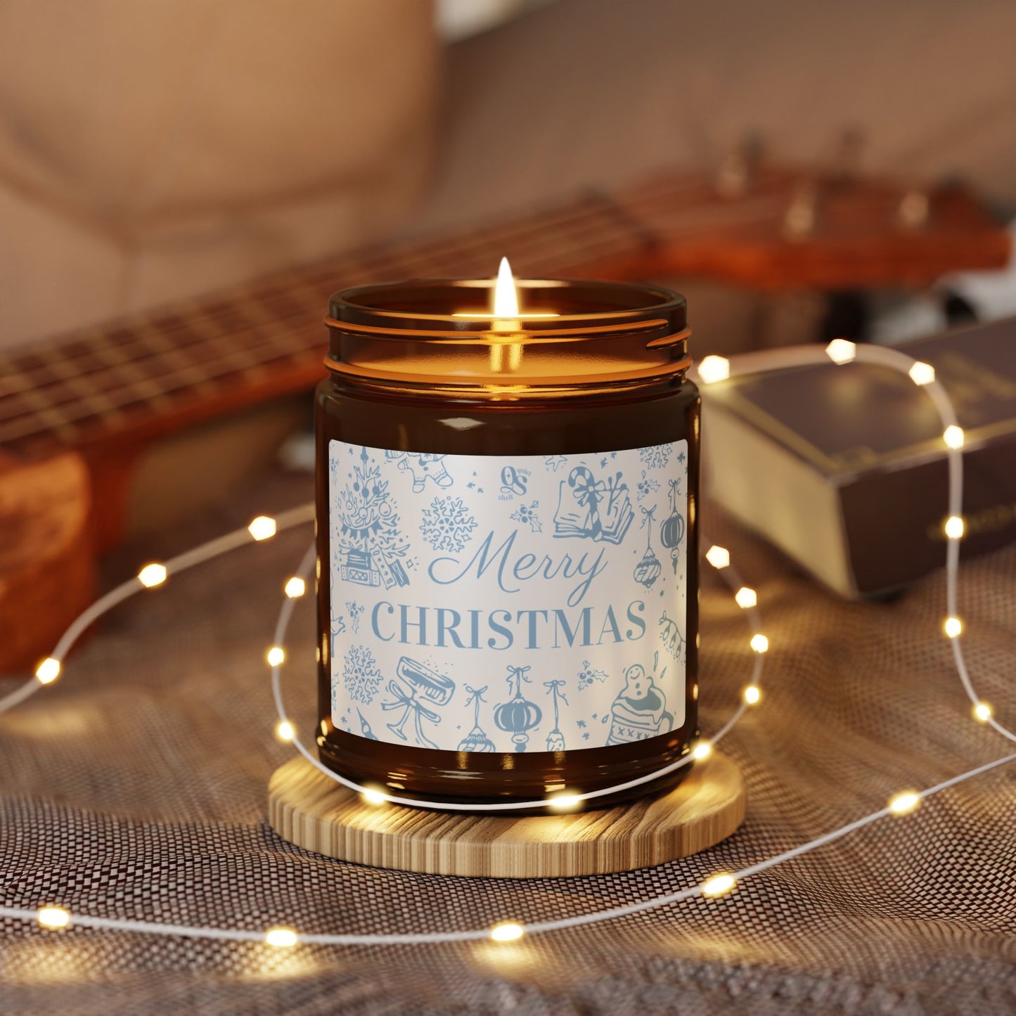 Candle with a 'Merry Christmas' label on a wooden coaster surrounded by string lights and a guitar in the background.