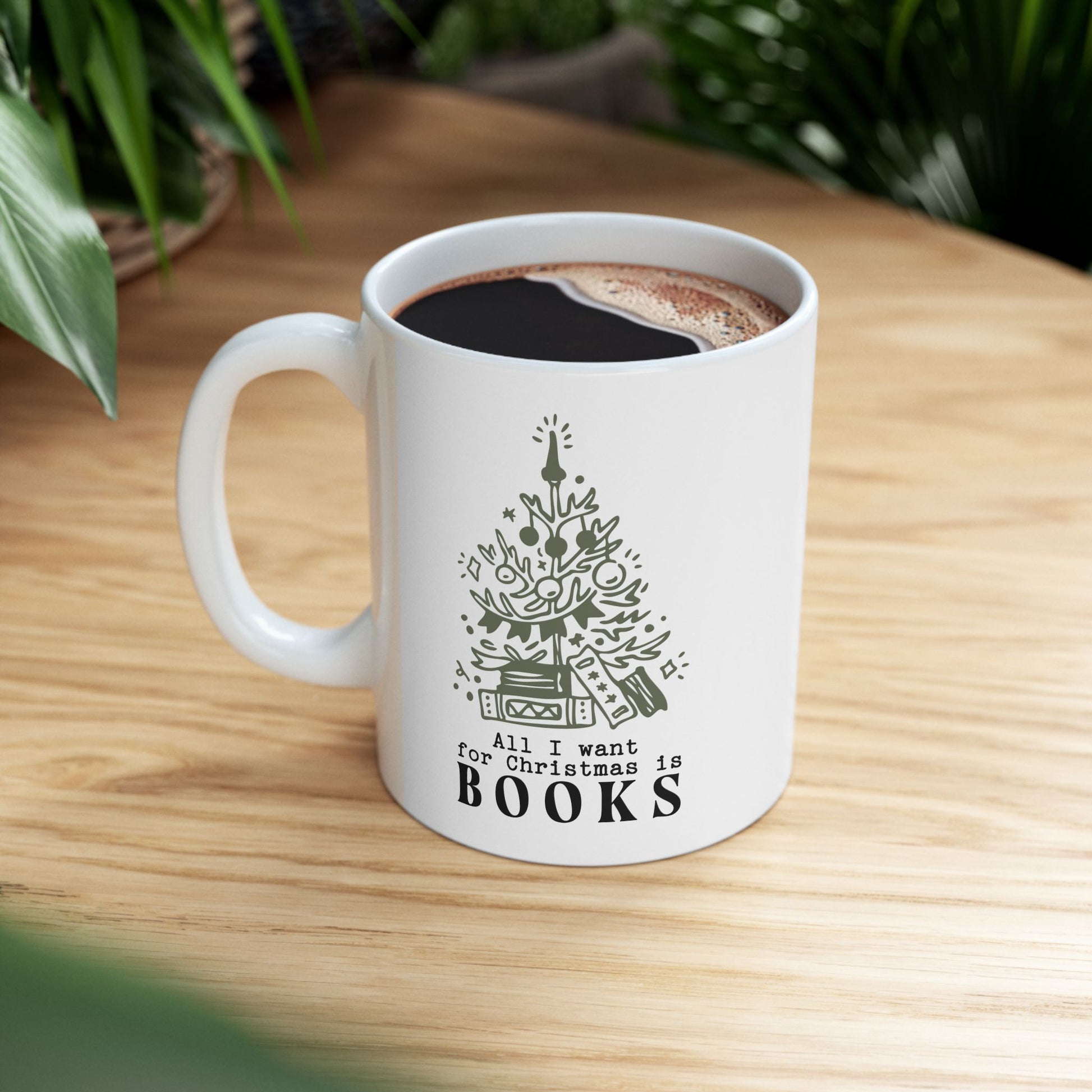 White mug with a Christmas tree design and 'All I want for Christmas is books' text on a wooden table.