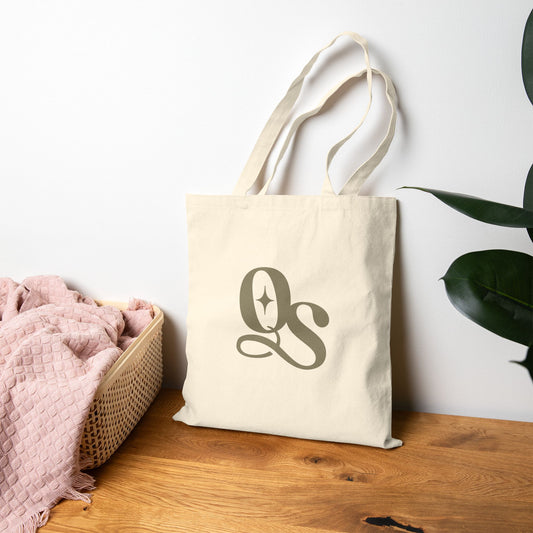 Beige tote bag with 'QS' logo on a wooden surface with a white wall and plant in the background