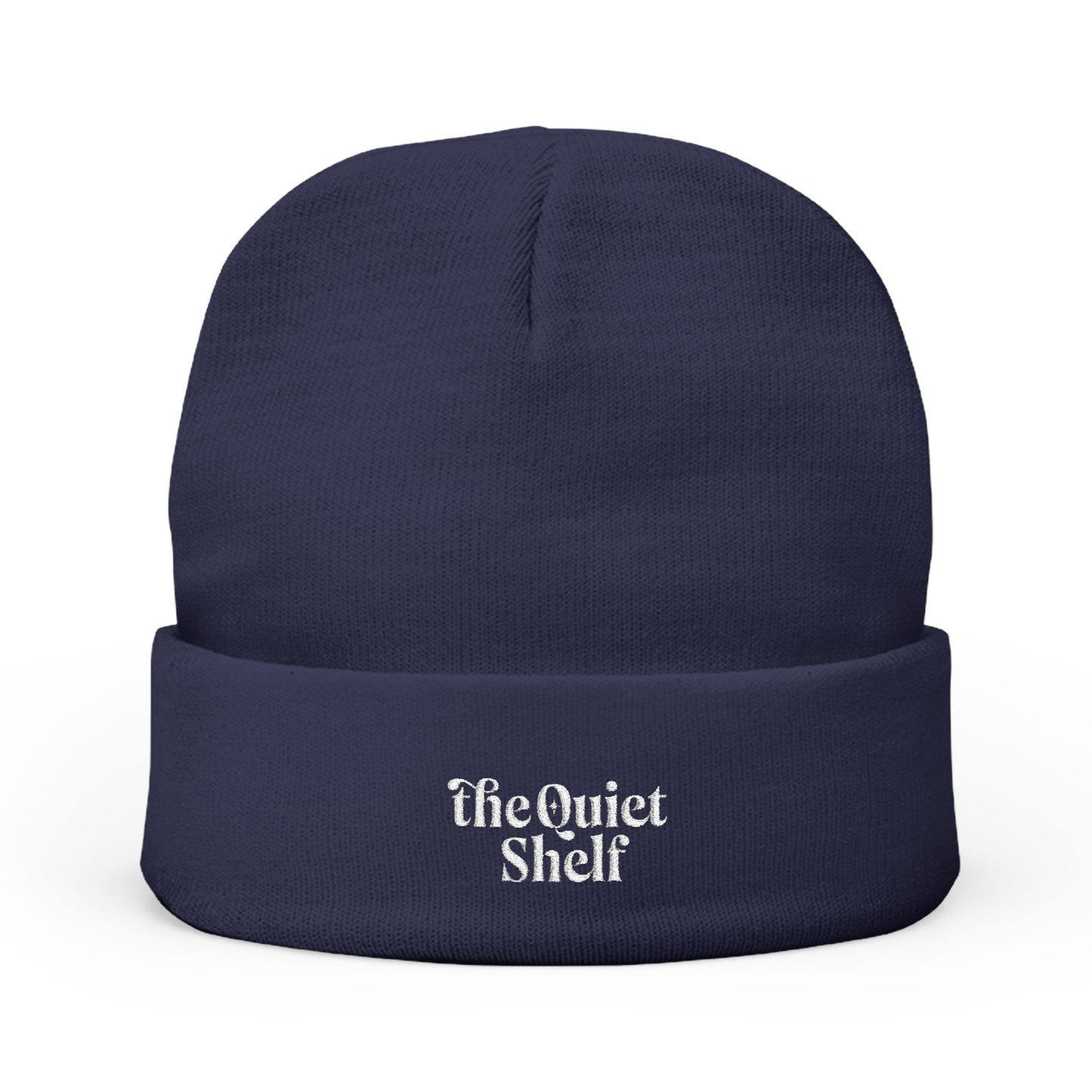 A navy blue cuffed beanie with 'The Quiet Shelf' embroidered in white on the fold-over cuff.
