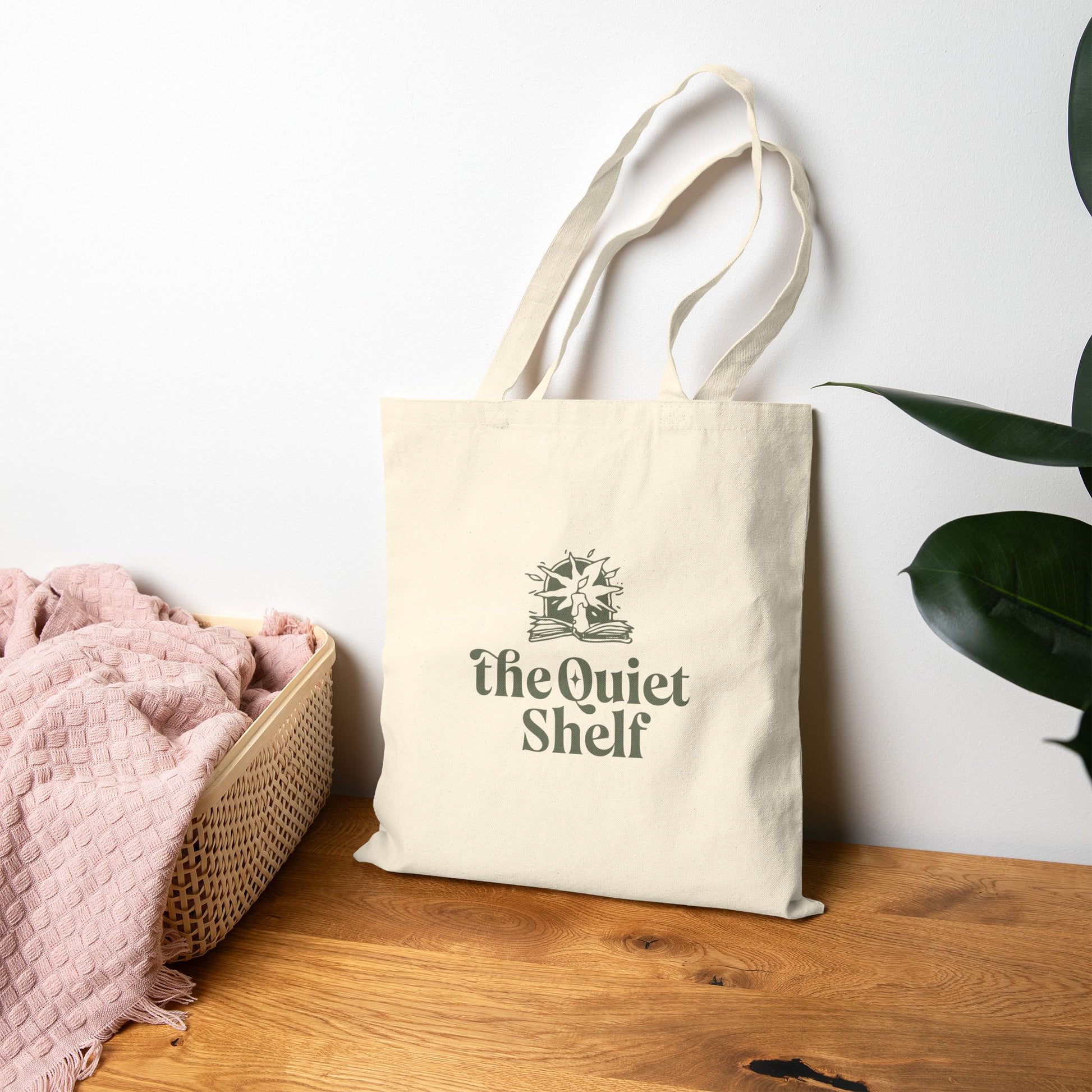 Beige tote bag with The Quiet Shelf logo on a wooden surface with a white wall and plant in the background.