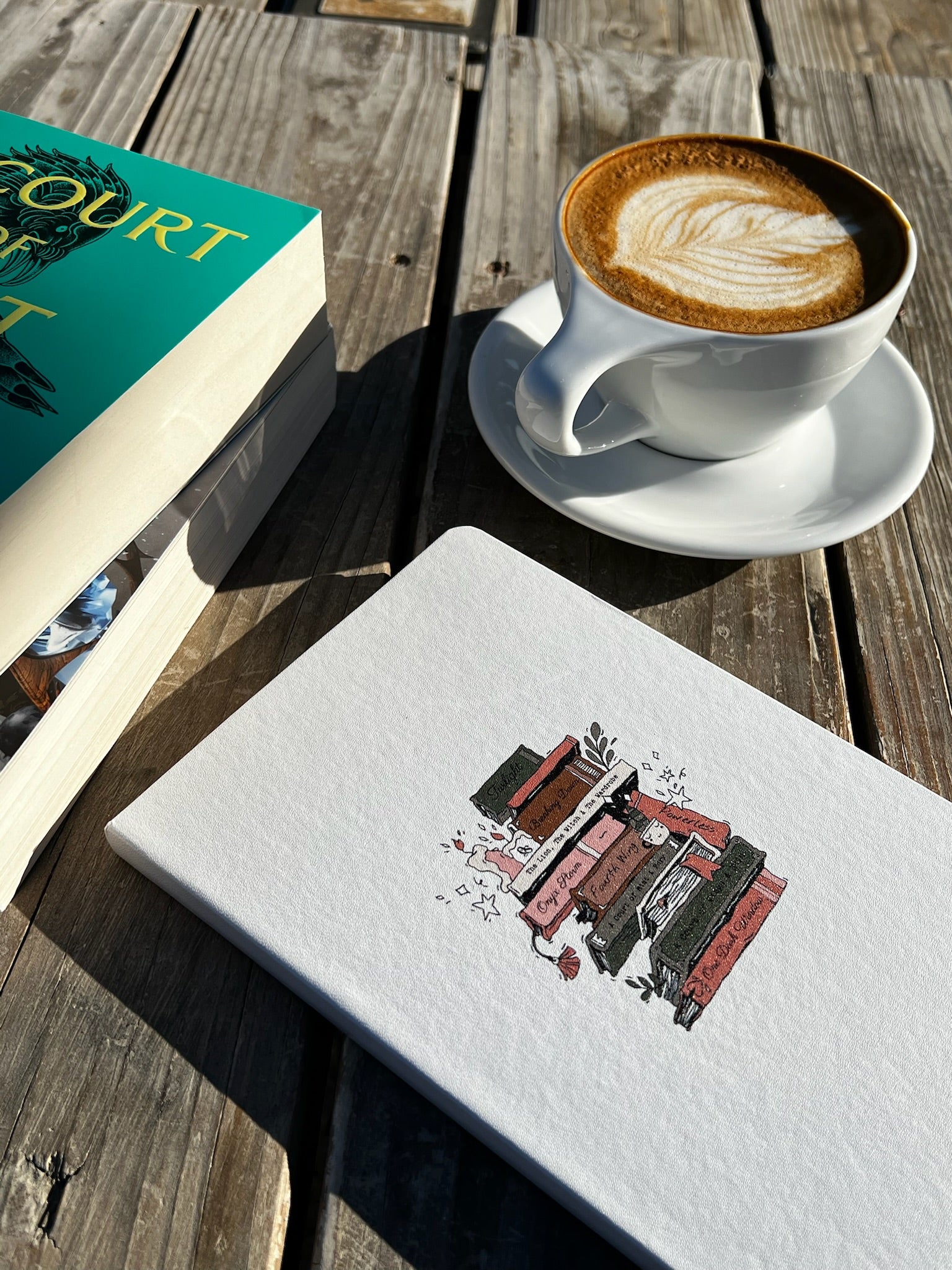 Cup of coffee with a saucer on a wooden table next to a book and a notebook with a design on the cover.