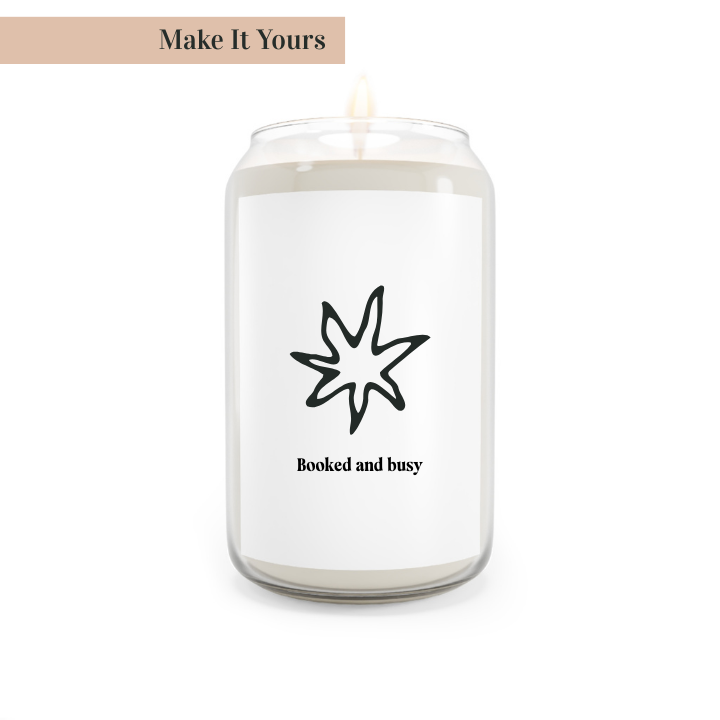 A white candle with a black star design and the text 'Booked and busy' on the label, with a lit wick, placed in a clear glass jar with a metal lid.