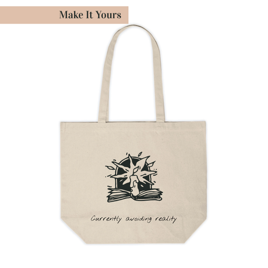 A beige canvas shopping tote with a black printed design featuring a book and the text 'Currently avoiding reality'. The tote has a flat bottom and two handles.