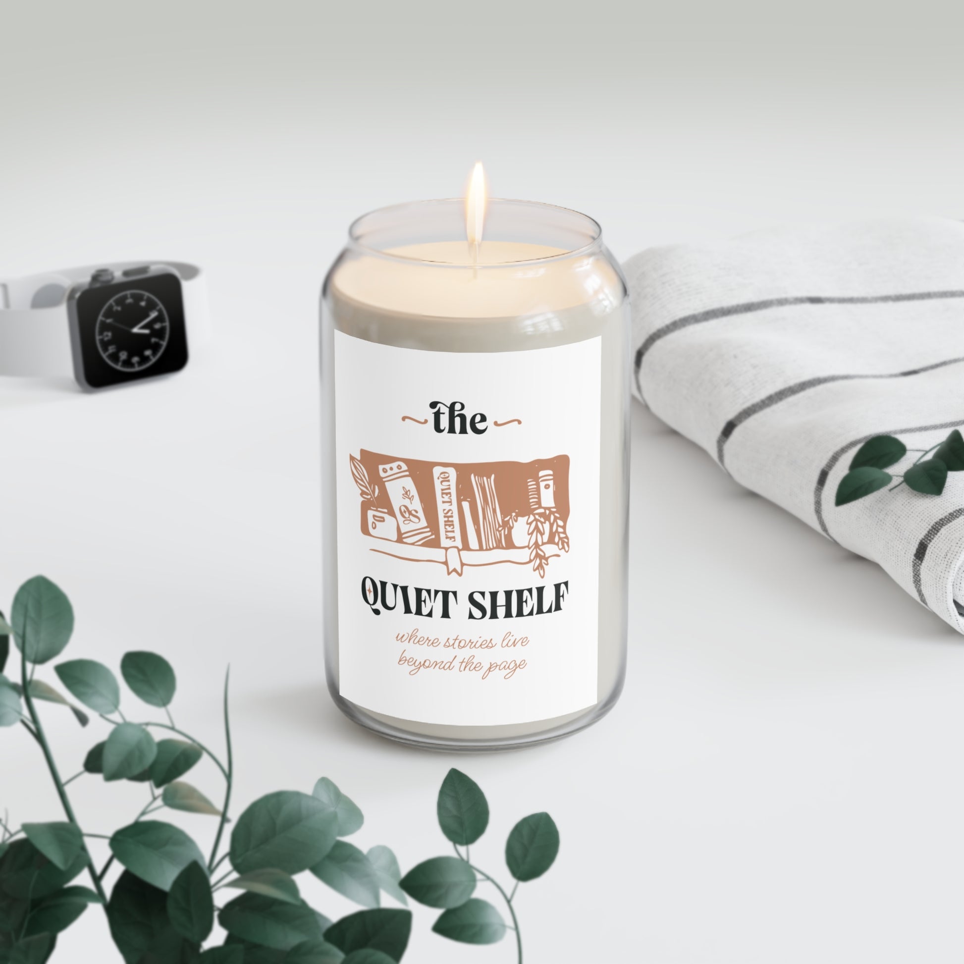 A soy candle in a clear glass jar with a minimalist label that features a book and the text 'the QUIET SHELF', with a lit wick, with a watch and a towel in the background and a plant in the foreground