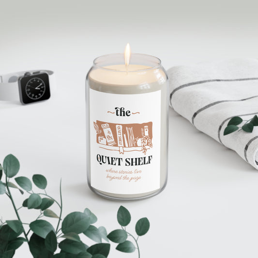 A soy candle in a clear glass jar with a minimalist label that features a book and the text 'the QUIET SHELF', with a lit wick, with a watch and a towel in the background and a plant in the foreground