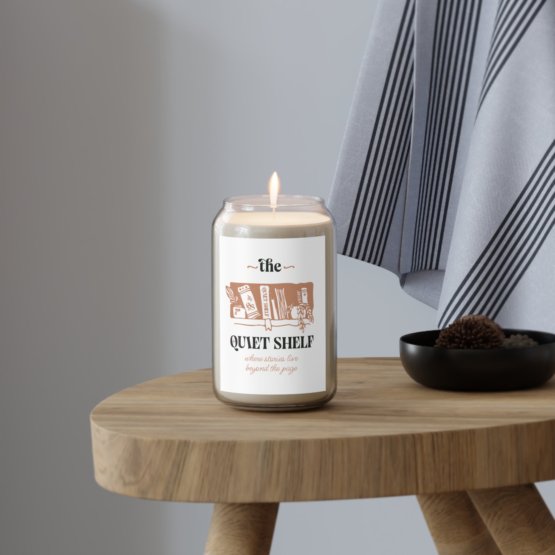 A soy candle in a clear glass jar with a minimalist label that features a book and the text 'the QUIET SHELF', with a lit wick, on a wood stool